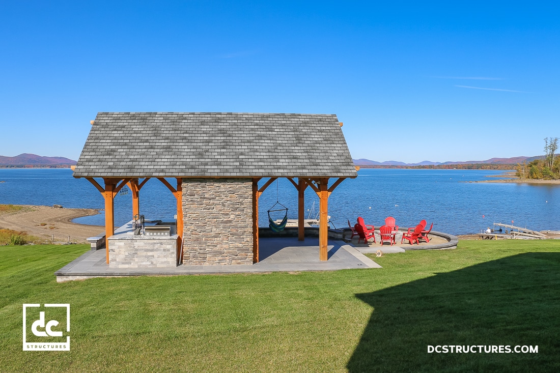 A lakeside pavilion with stone accents and wooden beams sits on grass. Behind it are a hammock and red chairs by a calm blue lake.