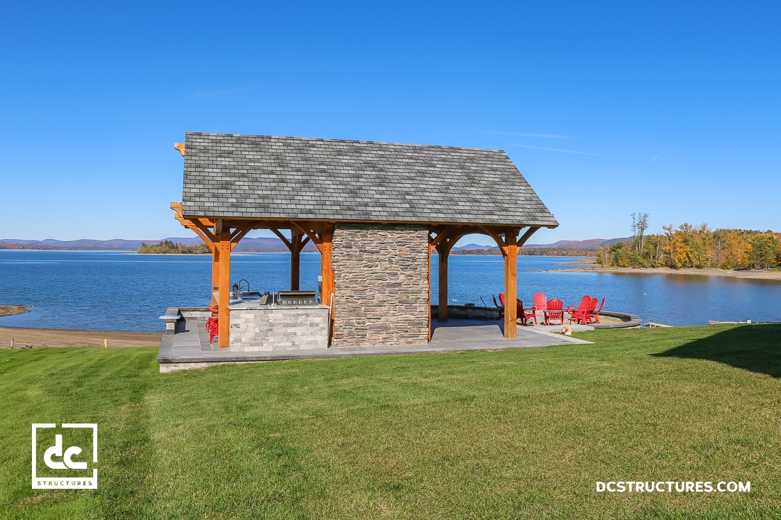 A lakeside pavilion with a stone fireplace and wooden beams sits on green grass near a calm blue lake. Red chairs are nearby.