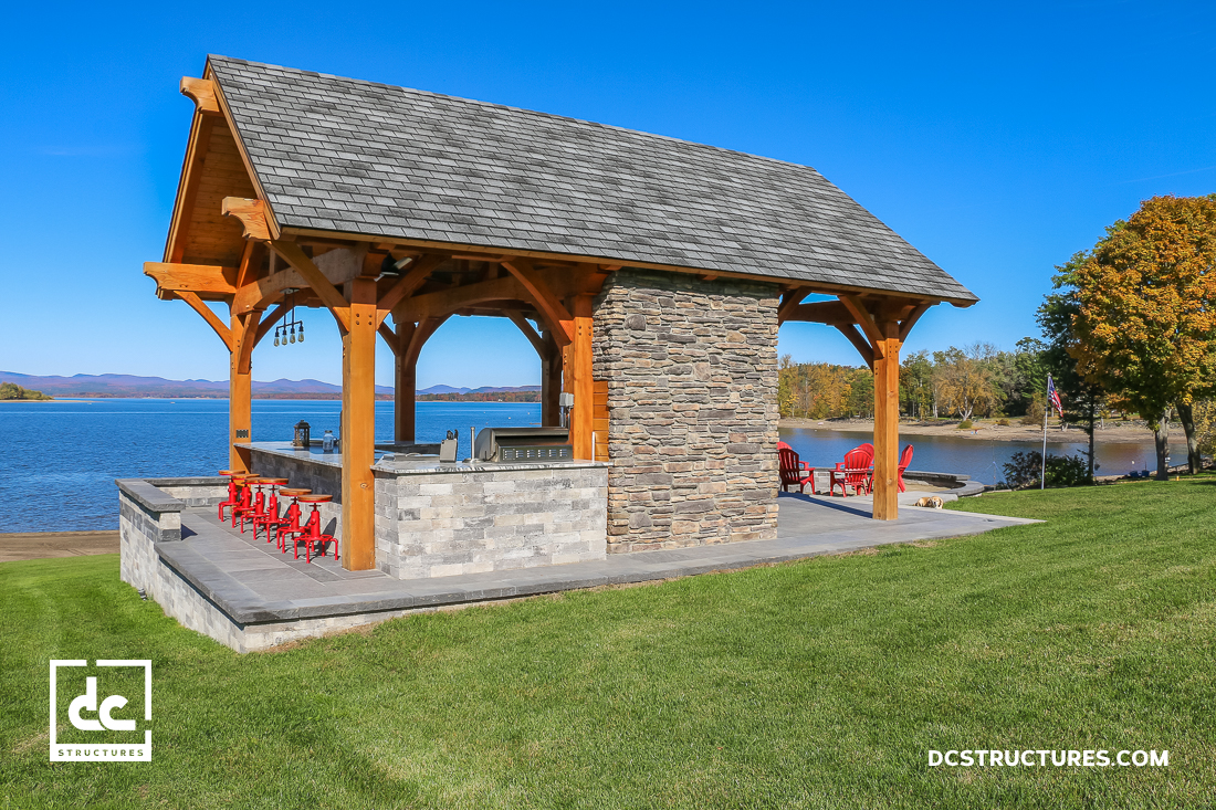 A wooden pavilion with a stone fireplace and bar seating overlooks a lake. Red chairs sit on a green lawn under a clear blue sky.