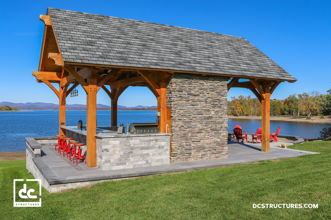 A lakeside outdoor pavilion with stone and wood structure, red bar stools, grill area, red Adirondack chairs, grass, lake, and mountains.