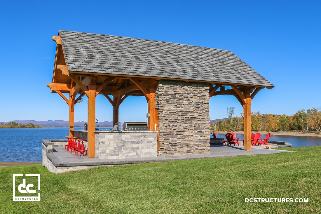 A lakeside pavilion with stone fireplace, wood beams, and patio seating on grass under clear sky. Red chairs by water, mountains beyond.