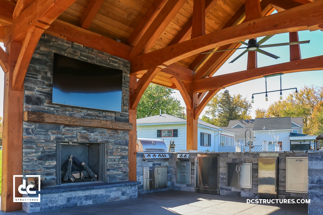 A covered outdoor kitchen features a stone fireplace with a mounted TV, grill, sink, and storage under a wood pavilion with fan.