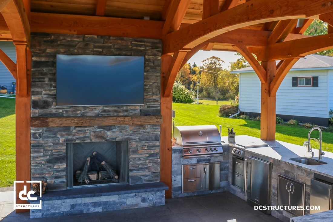 Covered outdoor kitchen with stone fireplace, mounted TV, stainless steel grill, sink, and cabinets under a wooden pergola. Green lawn and a small white house are visible in the background.
