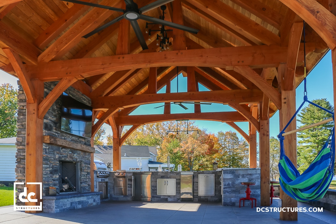 A covered outdoor kitchen and lounge with timber framing, stainless steel appliances, bar seating, a hammock, trees, and houses.