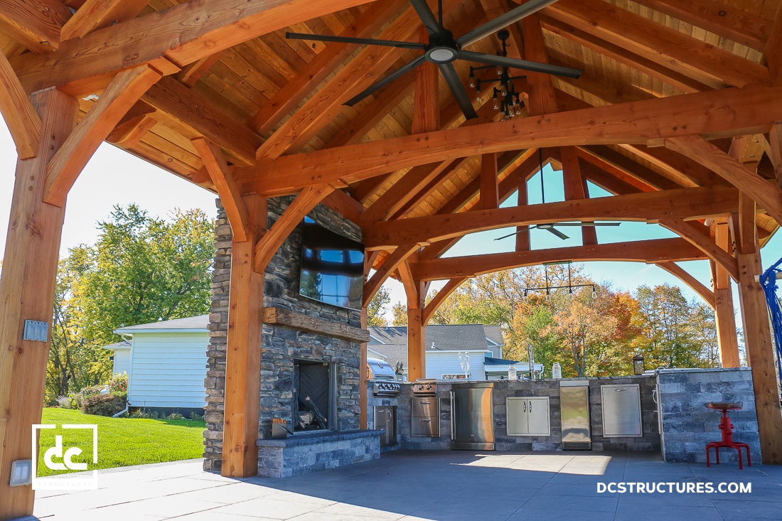 Spacious outdoor kitchen and living area under a wooden pavilion with exposed beams, stone fireplace, TV, appliances, ceiling fan, trees.