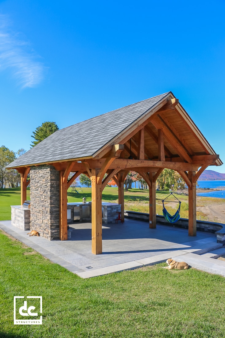 A wooden pavilion with a stone pillar stands on a paved area near a lawn and beach, with tables, a hammock, grill, and two dogs nearby.