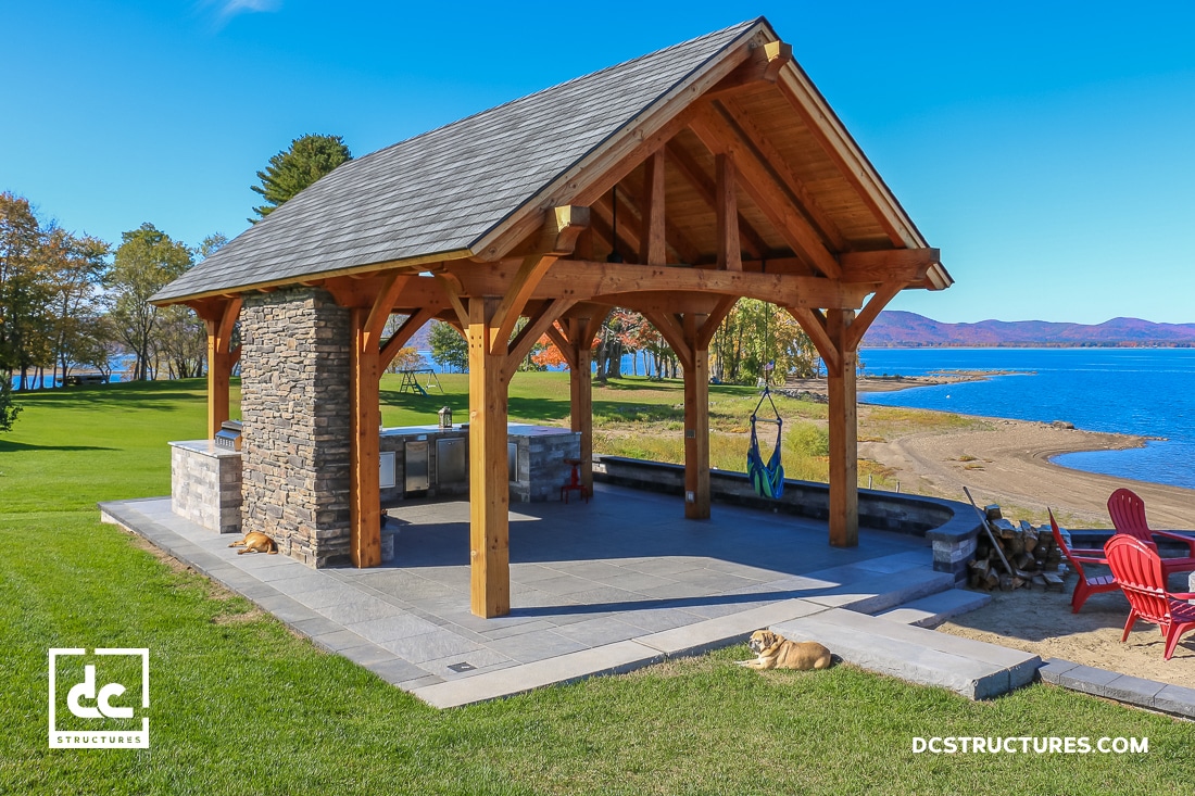 A wooden pavilion with a stone grill sits on a paved patio by a lake. Red chairs face the water, and two dogs rest nearby. Trees and green grass surround the area under a clear blue sky.