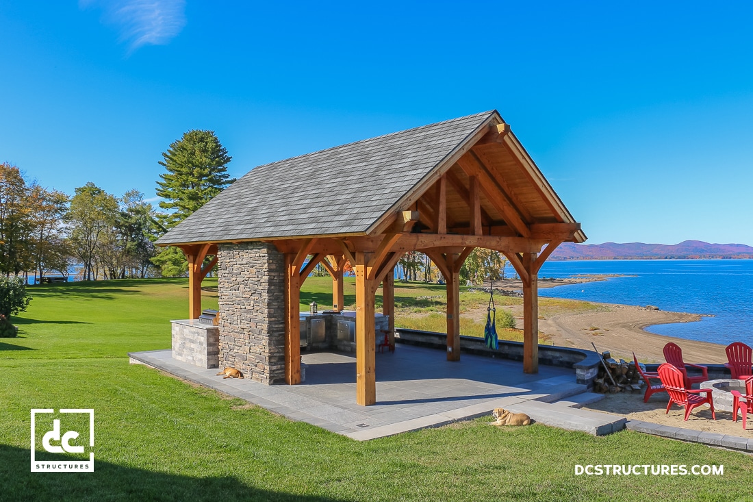 A wooden pavilion with a stone fireplace sits on a grassy lawn by a sandy beach and blue lake, red chairs nearby, trees behind.