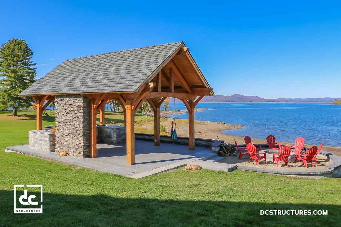 A wooden pavilion with a stone fireplace stands by a sandy lakeshore, red Adirondack chairs around a fire pit, mountains beyond.