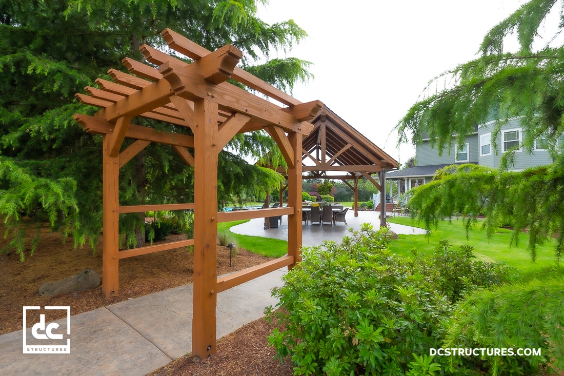 A wooden pergola stands on a landscaped path leading to a covered patio with chairs and a table, surrounded by green trees.