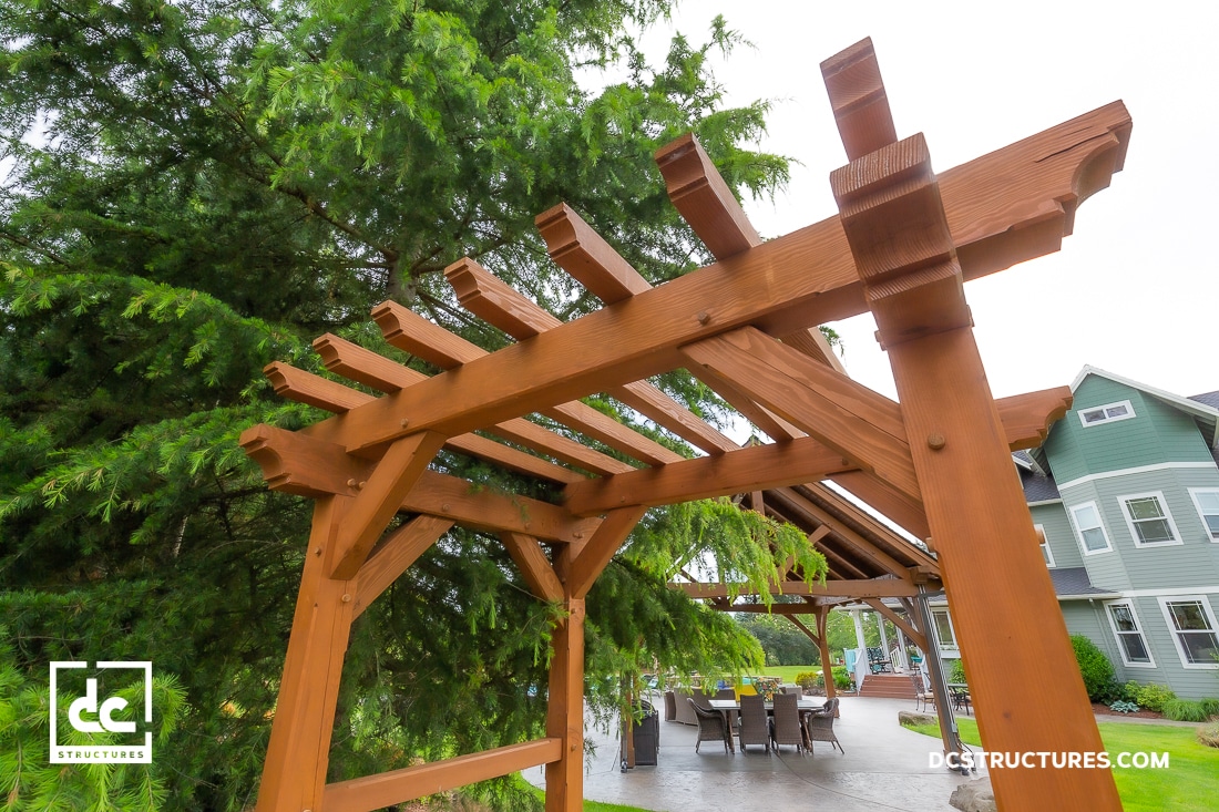 A close-up view of a wooden pergola in a backyard, with patio furniture and a green house in the background; lush trees surround the area.