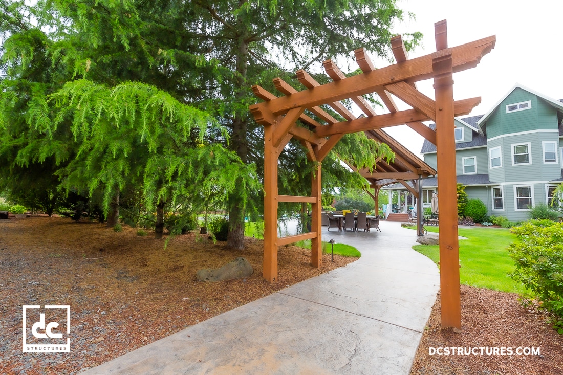 A wooden pergola stands over a curved concrete pathway leading to a green house with white trim, surrounded by trees and landscaping.