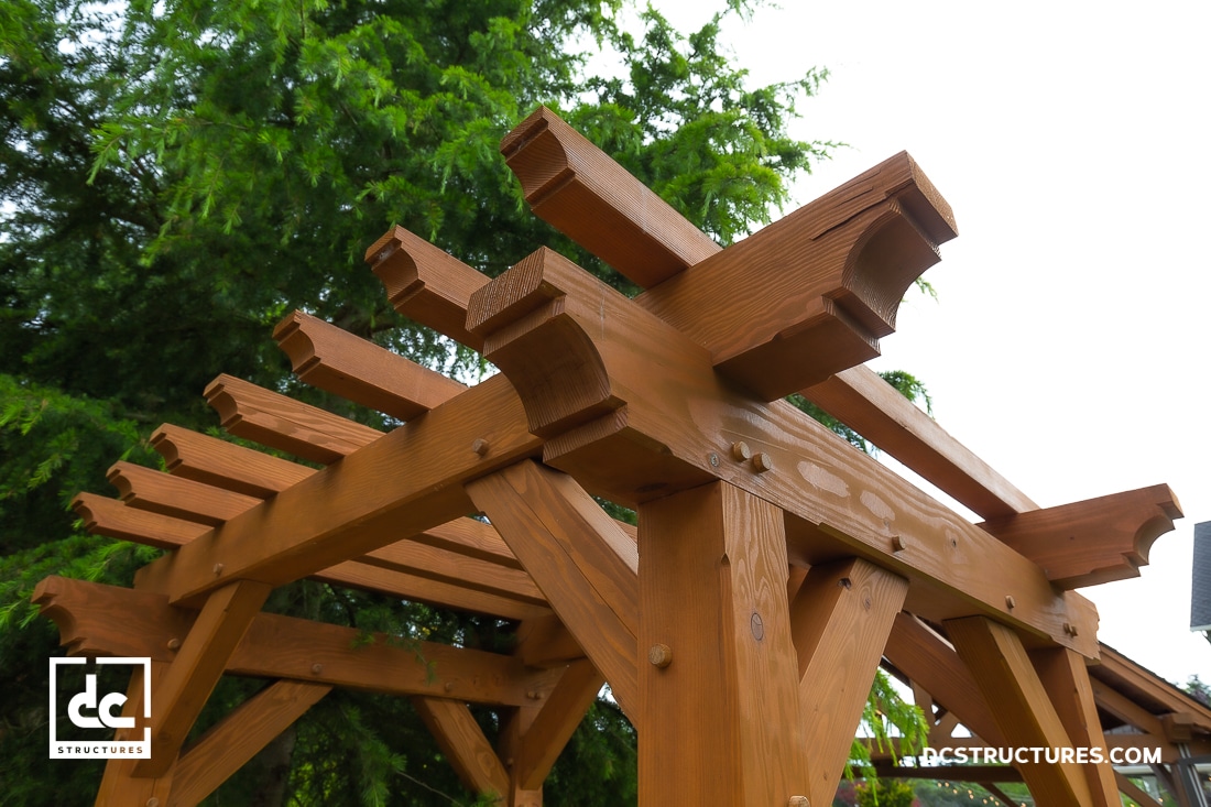 Close-up of a wooden pergola structure with decorative beams and joints, set against a background of green trees.