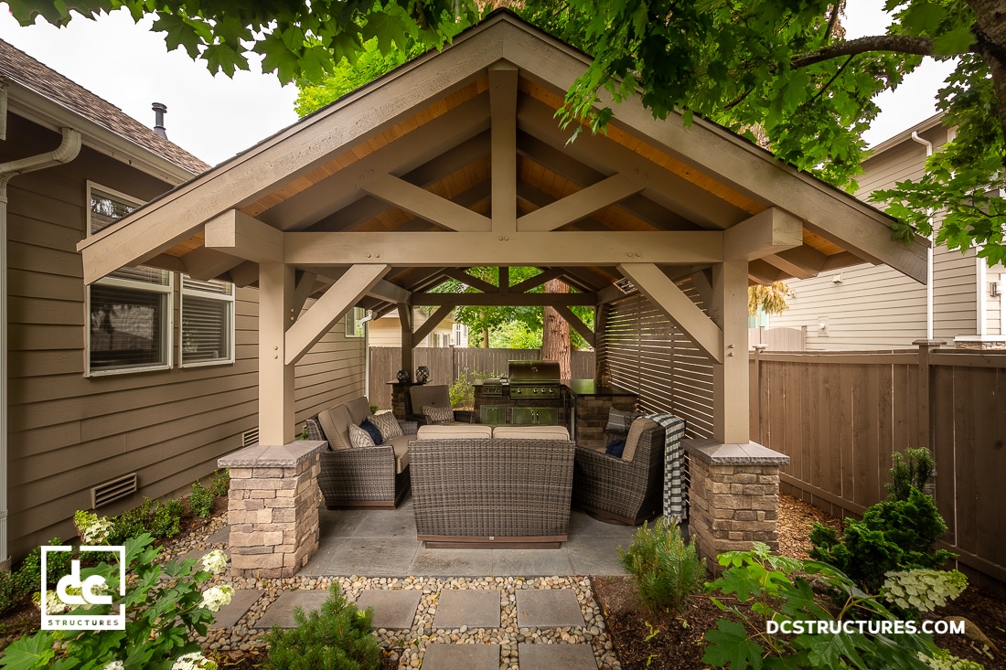 A covered outdoor patio with wicker furniture, stone pillars, and a wooden gazebo roof sits between two houses amid greenery.