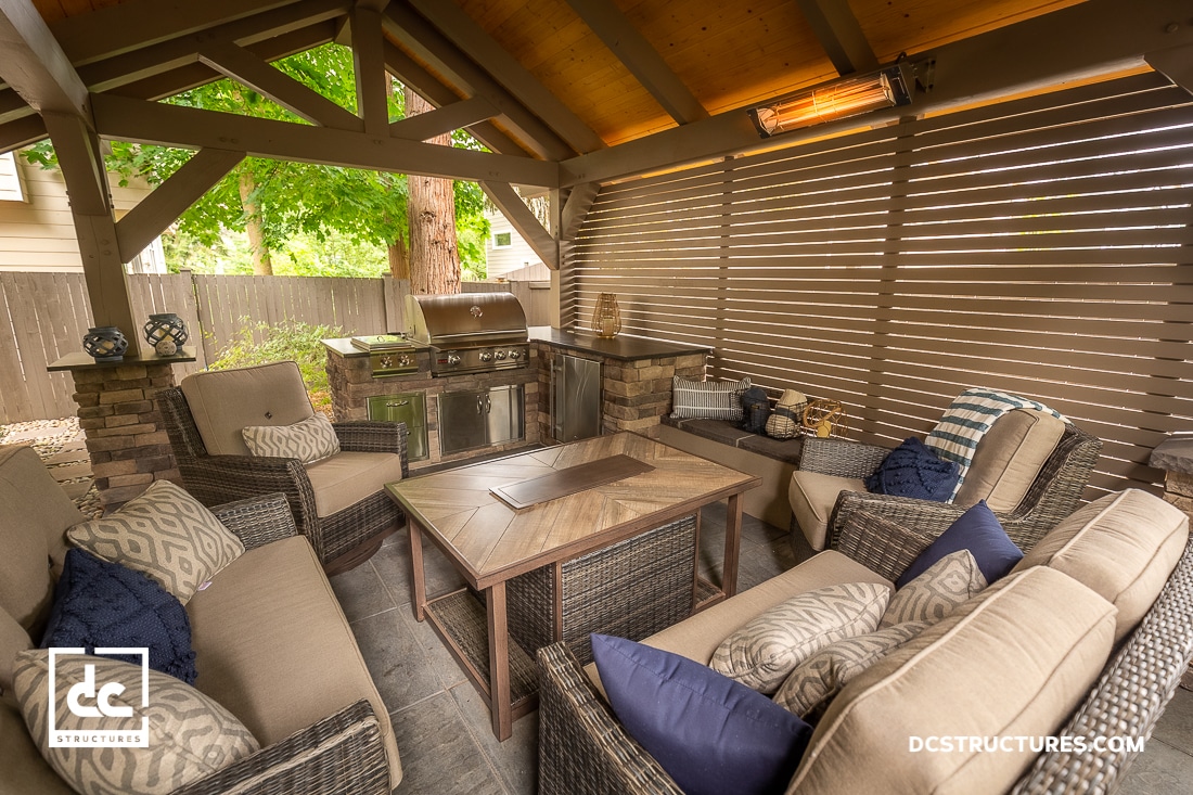 A cozy outdoor patio with cushioned wicker furniture, a coffee table, a grill, and a wooden pergola surrounded by greenery.
