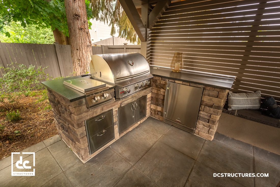 Outdoor kitchen with built-in stainless steel grill, storage cabinets, and stone countertops under a covered patio. Wooden slatted wall and trees provide a natural, shaded setting.