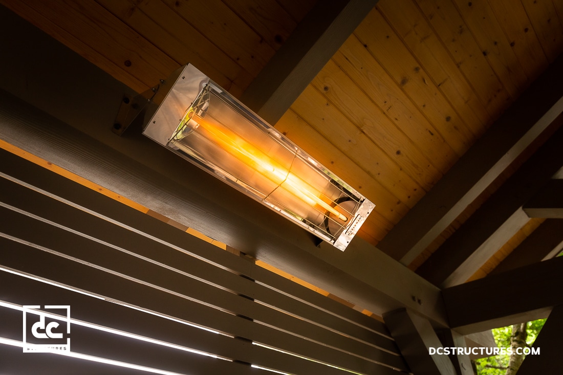 A close-up view of an outdoor mounted electric heater glowing orange, attached to a wooden ceiling with beams.