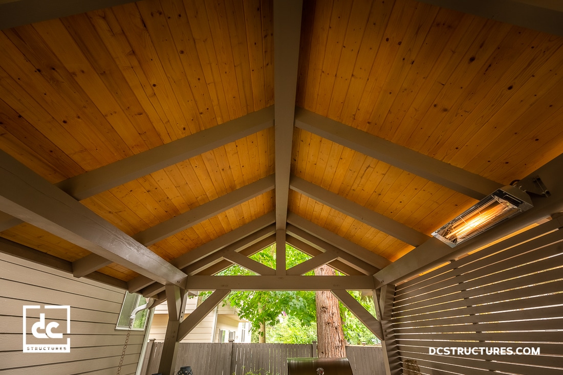 Wooden patio cover with exposed beams and a natural wood ceiling, partially enclosed by slatted walls. Electric heater on right. Trees and fence seen through open end.