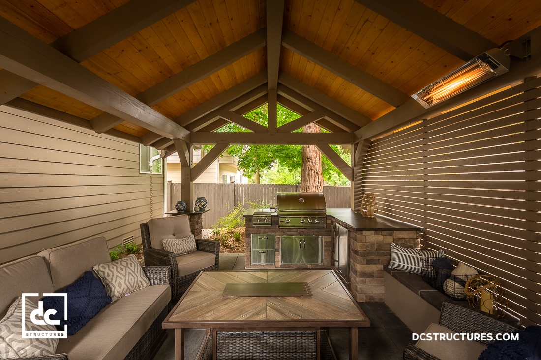 Covered outdoor patio with wooden beams, wicker furniture, modern grill, heating lamp, cozy seating area, outdoor kitchen, greenery.