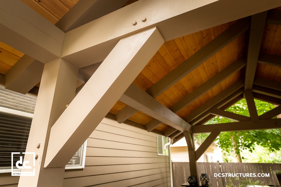 A close-up view of timber framing with beige beams and a wooden ceiling under a covered outdoor structure, adjacent to a house.