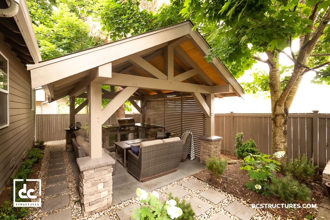 A covered outdoor patio with cushioned wicker furniture sits under a wooden pavilion, surrounded by a fenced yard with greenery.