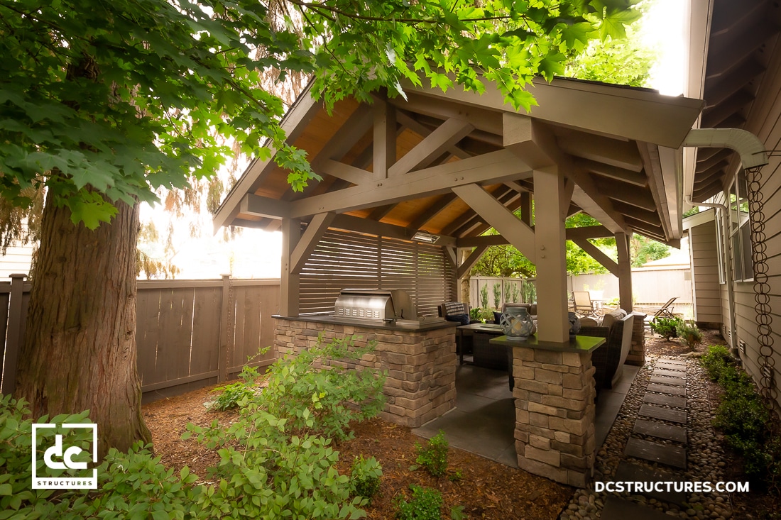 A covered outdoor patio with a wooden and stone structure, built-in grill, countertop, seating area, greenery, and wooden fence.
