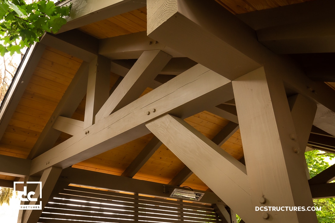 Close-up view of a wooden pergola’s roof structure, showing tan and brown beams with visible bolts and a slatted roof design. Sunlight and green leaves appear in the corners.