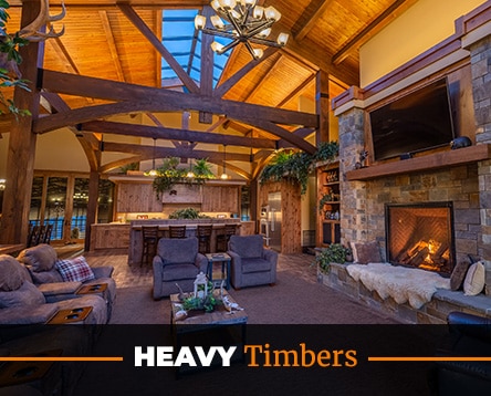 A rustic living room with a high wooden ceiling and beams, reminiscent of barn home kits, features a stone fireplace, plush sofas, and a large window. The room is warmly lit, and there's a banner across the bottom that says "Heavy Timbers.