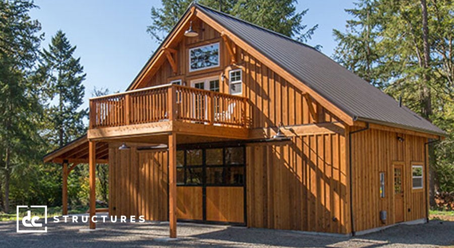 A rustic two-story wooden cabin with a sloped metal roof. It features a spacious balcony and large garage doors. Surrounded by trees and clear skies, the cabin is set on a gravel surface. A logo reads "DC Structures" in the lower left corner.