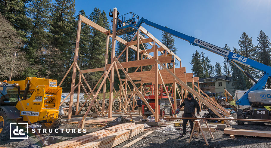 Construction site with workers assembling a wooden structure using cranes and machinery. Tall trees and a house are visible in the background. The scene is busy with various construction materials and equipment scattered around.