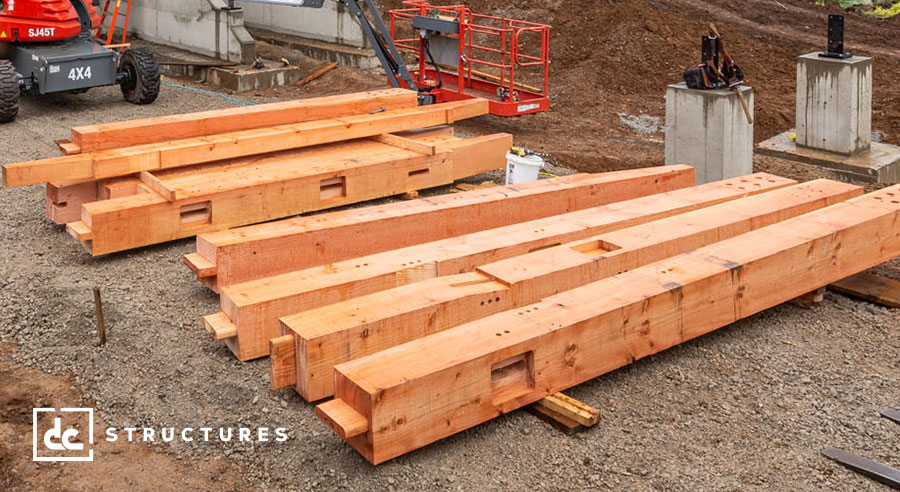 Stacks of large wooden beams lie on the ground at a construction site. Nearby, construction equipment and materials are visible, including a red lift and concrete bases. The site appears to be in early stages of building.