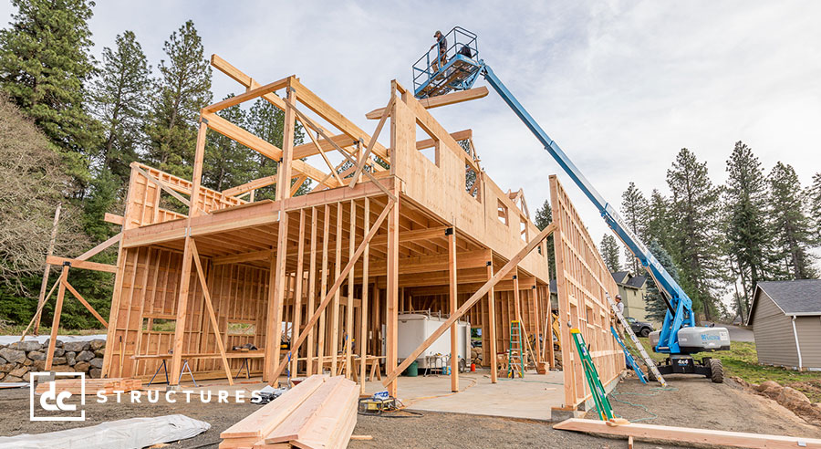 A partially constructed wooden house in a forested area. A blue crane lifts a worker on a platform to the upper level. Construction materials and tools are scattered around the site. The sky is overcast.