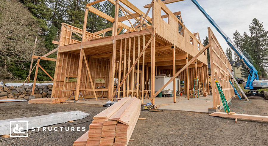 A wooden frame of a house under construction, with walls partially erected. Various construction materials and tools are scattered around, and a crane assists in assembling the structure. Trees are visible in the background.