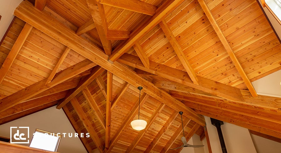 A ceiling with exposed wooden beams forming a geometric pattern. The wood has a warm finish, and there are two ceiling fans. Natural light filters through a small window.