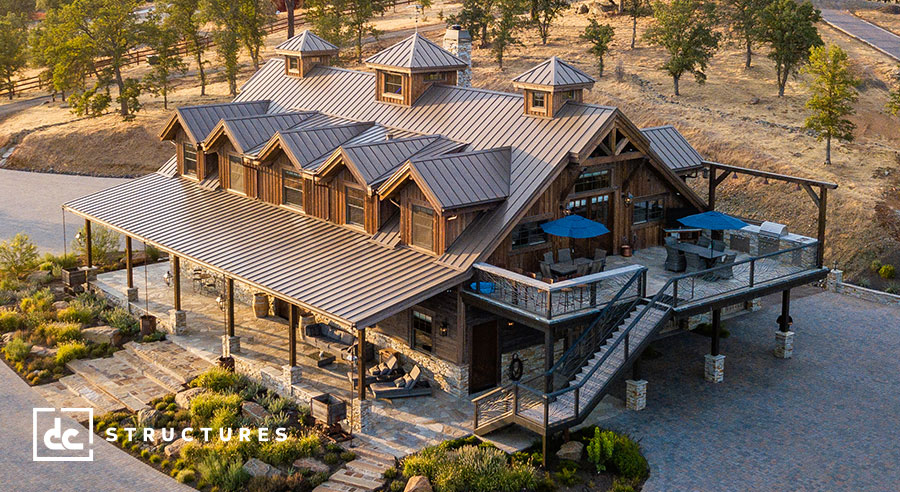 Aerial view of a large, rustic wooden building with multiple peaked roofs and wide porches. The structure is surrounded by a landscaped area with trees and shrubs. A deck with tables and blue umbrellas is visible on one side.