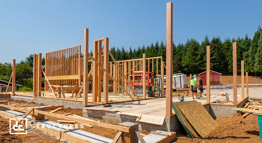 Construction site with wooden frames of a building in progress. Workers are seen on the partially built structure, surrounded by tools and materials. A red shed and trees are in the background under a clear blue sky.