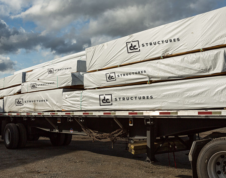 A flatbed truck carries large, wrapped stacks of construction materials labeled "DC Structures." The packages are secured and the scene is set under a partly cloudy sky.