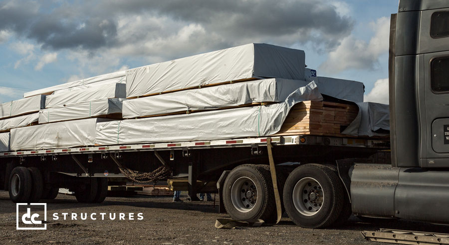 A flatbed truck loaded with stacks of lumber wrapped in protective covers is parked on a construction site. The sky is partly cloudy in the background. The logo "dc structures" is in the bottom left corner.