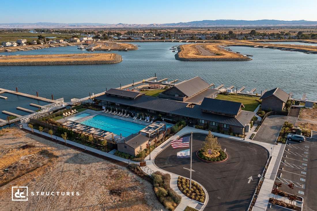 Aerial view of a large waterfront property with modern buildings and a swimming pool. The complex is surrounded by water and docks. An American flag waves in front, and the landscape includes dry vegetation and adjacent parking.