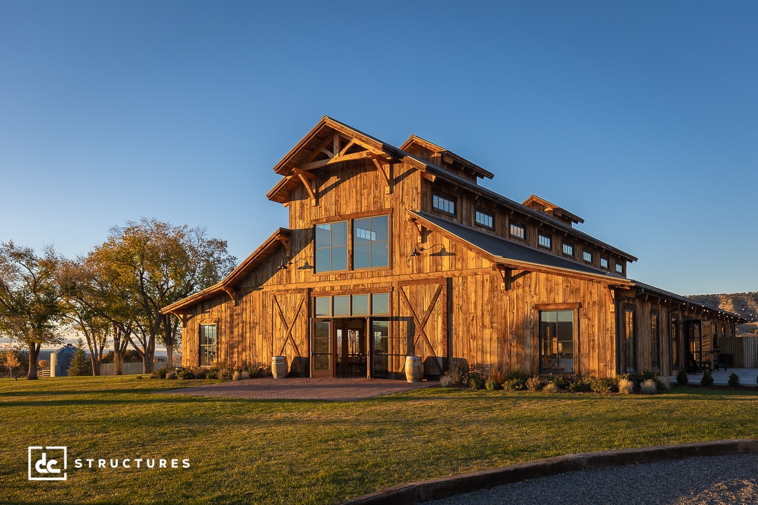 A large, rustic wooden barn-style building with multiple windows and a spacious entrance stands under a clear blue sky. Surrounded by green grass and trees, the structure is well-lit by the warm glow of the setting sun.