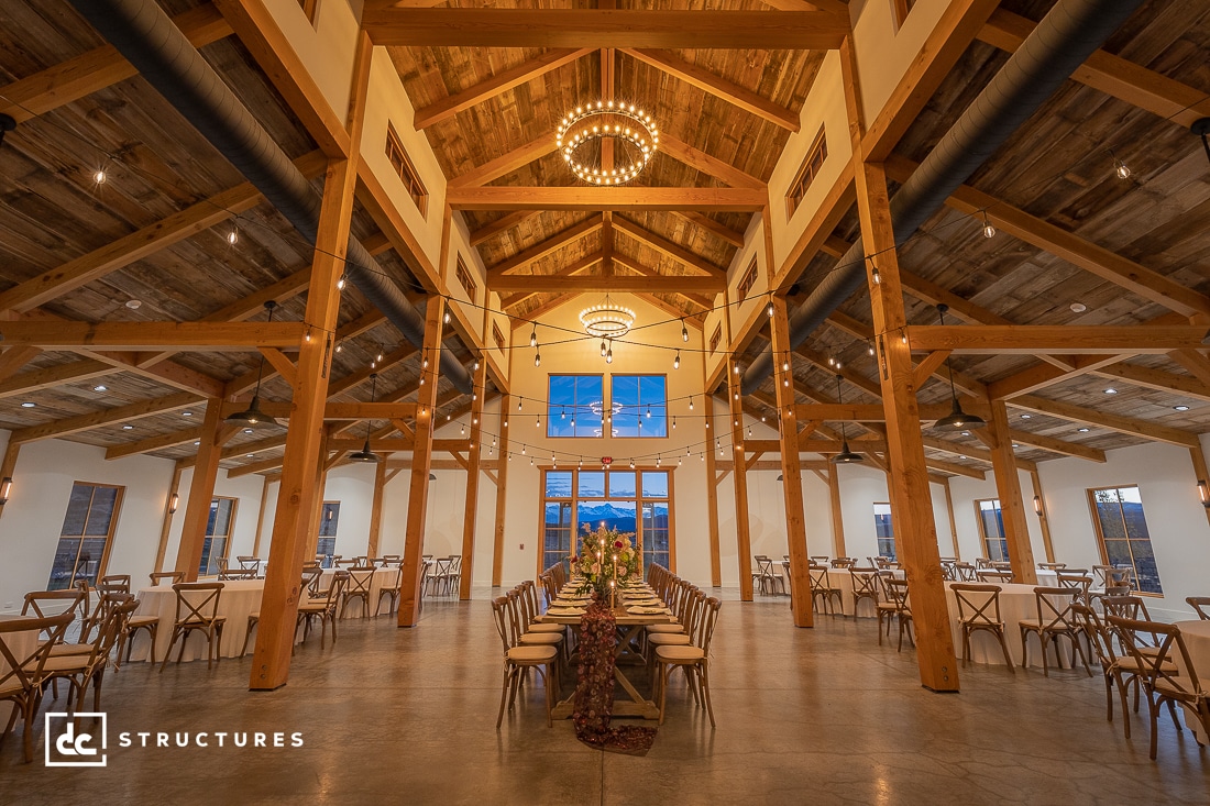 A spacious rustic event hall with a high wooden ceiling and warm lighting. Long tables with white tablecloths and elegant decorations are set for a banquet. Wooden beams and chandeliers enhance the cozy, inviting atmosphere.