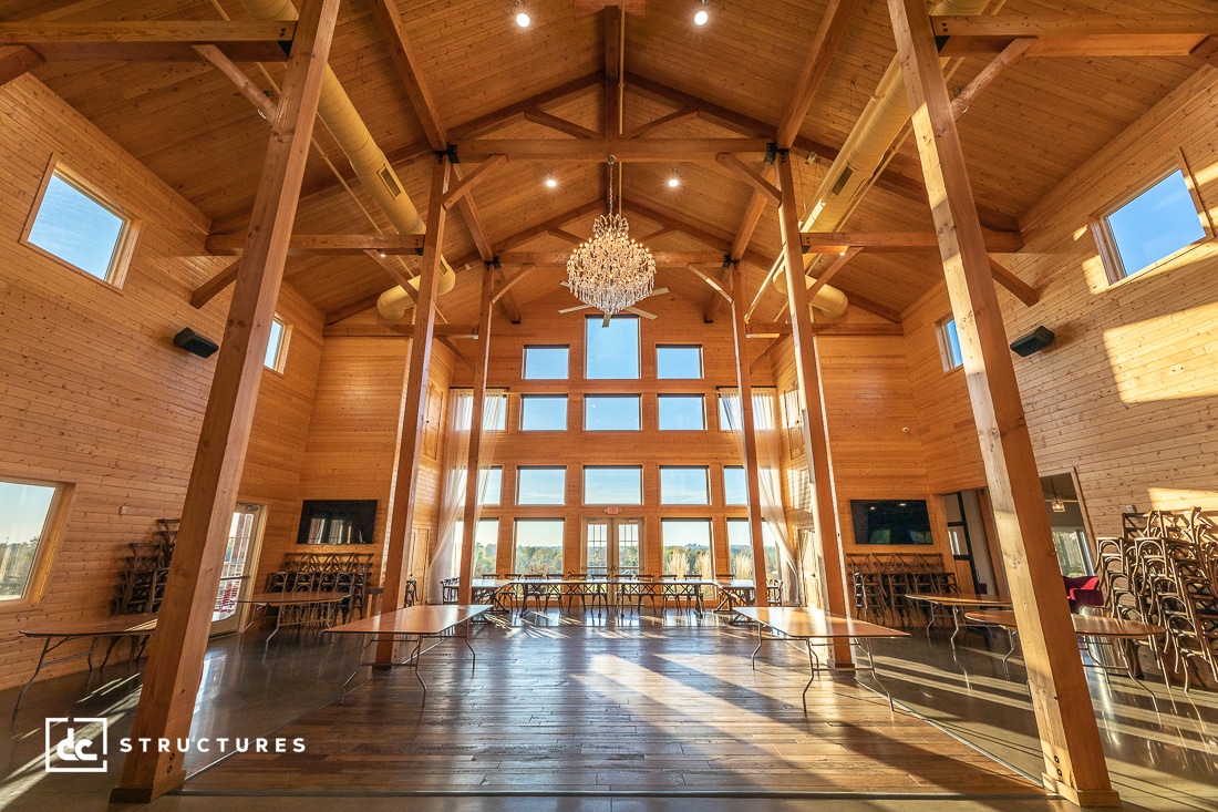 Spacious wooden hall with high ceilings and large windows. A chandelier hangs at the center. Tables are set around the room, and chairs are stacked in the corner. Sunlight streams in, highlighting the woodwork. Logo in the corner reads “DC Structures.”.