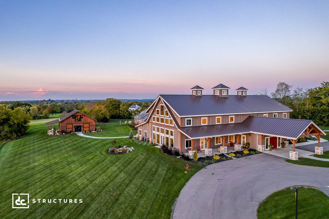 A large, modern barn-style building with multiple gables and a metal roof stands on a manicured lawn. A smaller barn is visible in the distance, surrounded by trees under a clear sky at sunset. A winding driveway leads to the main structure.