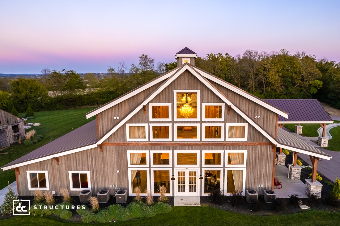 A large, rustic barn-style building with wooden siding, multiple windows, and a cupola on top. The surrounding area has lush green grass and trees, with a clear evening sky in the background. A logo in the corner reads "DC Structures.