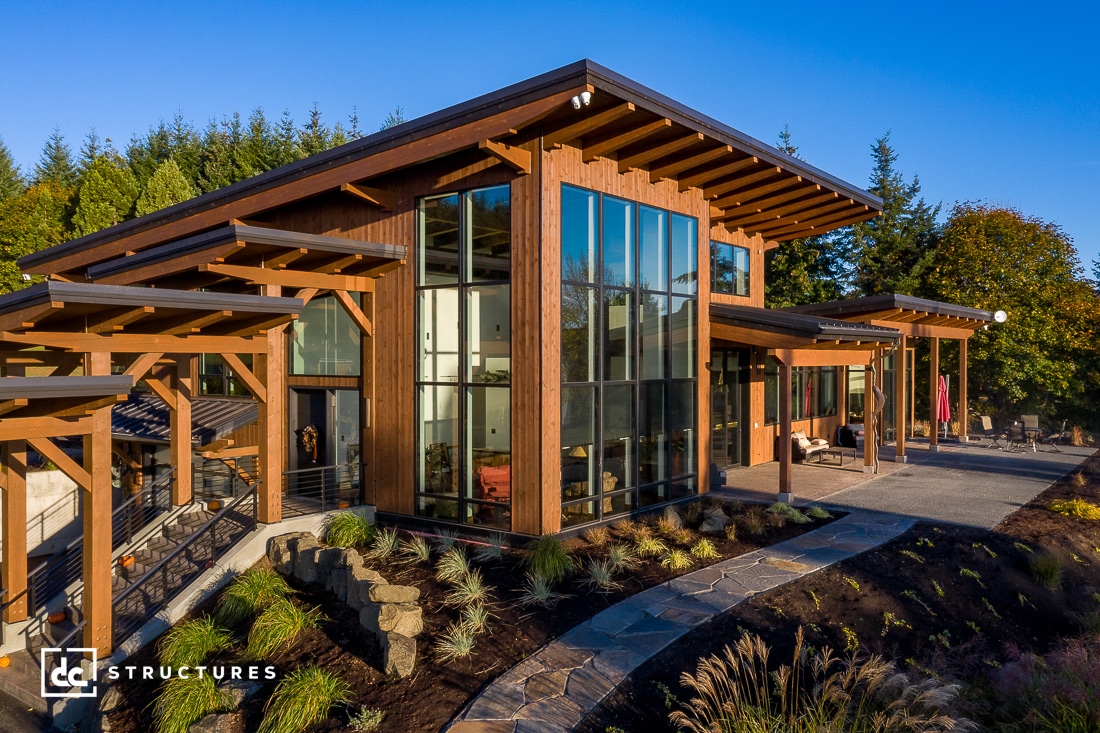 Modern two-story house with large glass windows and wooden accents surrounded by a landscaped garden. A stone pathway leads to the entrance. The structure features multiple overhangs and a mix of horizontal and vertical lines, giving it a contemporary look.