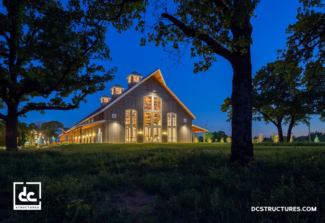 A large barn-style building with illuminated windows, featuring a custom design offering, sits on a grassy lawn surrounded by trees at dusk. The deep blue sky enhances the structure, beautifully highlighted by exterior lights.