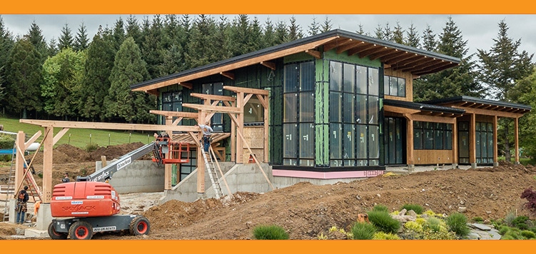 A modern house under construction, featuring a custom design offering with large glass windows and wooden beams. Surrounding the structure are piles of earth and construction equipment. In the background, tall trees sway beneath a cloudy sky.