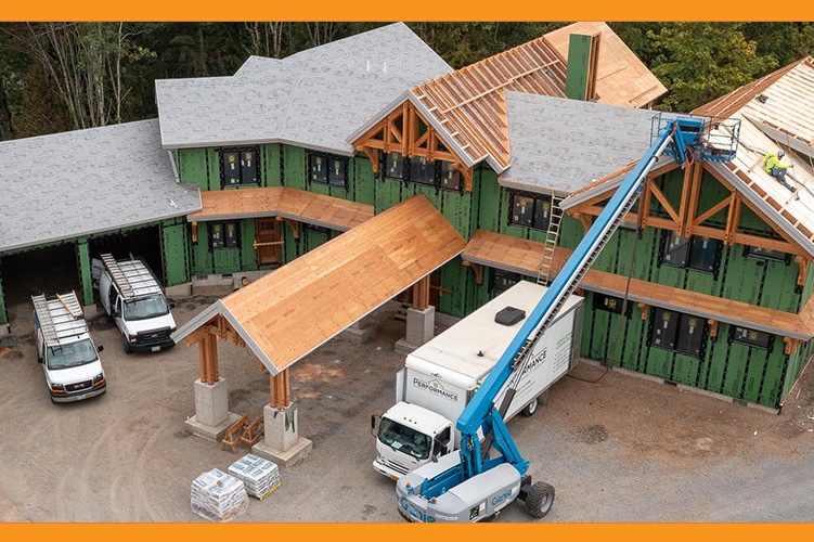 Aerial view of a large, custom-designed house under construction, featuring green exterior walls and wooden roofing. Two vans are parked nearby, along with a truck. A worker on a blue hydraulic lift is carefully installing roofing panels. Trees and dirt surround the site.