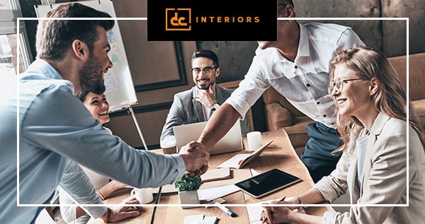 A group of colleagues in a modern office setting are smiling and engaged in discussion. Two people are shaking hands across a table with laptops, notebooks, and a plant. A DC Interiors logo is at the top.