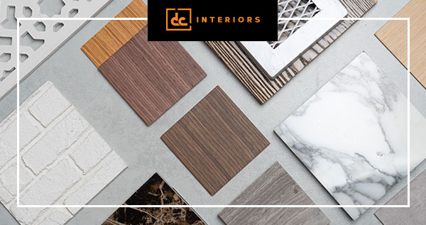 A variety of decorative material samples are arranged on a gray surface, featuring different textures and patterns such as wood, marble, and brick. A logo with the text "dc INTERIORS" is visible at the top of the image.